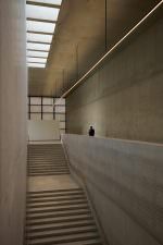 Tall concrete stairwell under a skylight, a thin line of light running diagonally; a lone man at the upper ledge, dwarfed by the space.