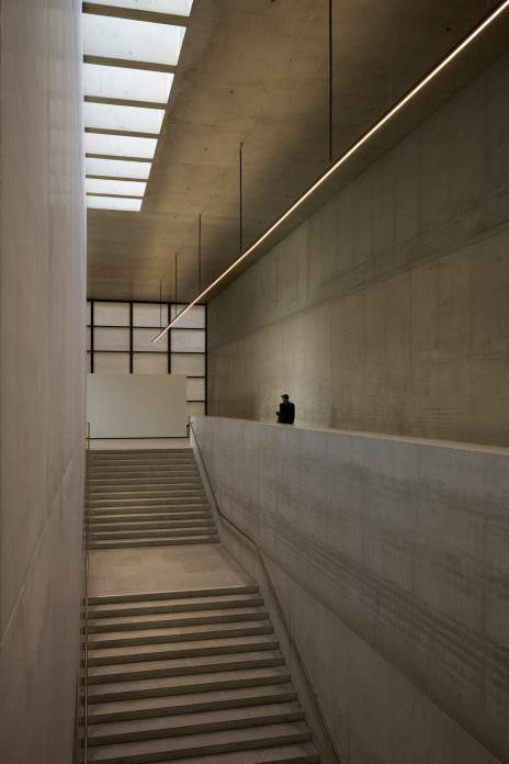Tall concrete stairwell under a skylight, a thin line of light running diagonally; a lone man at the upper ledge, dwarfed by the space.