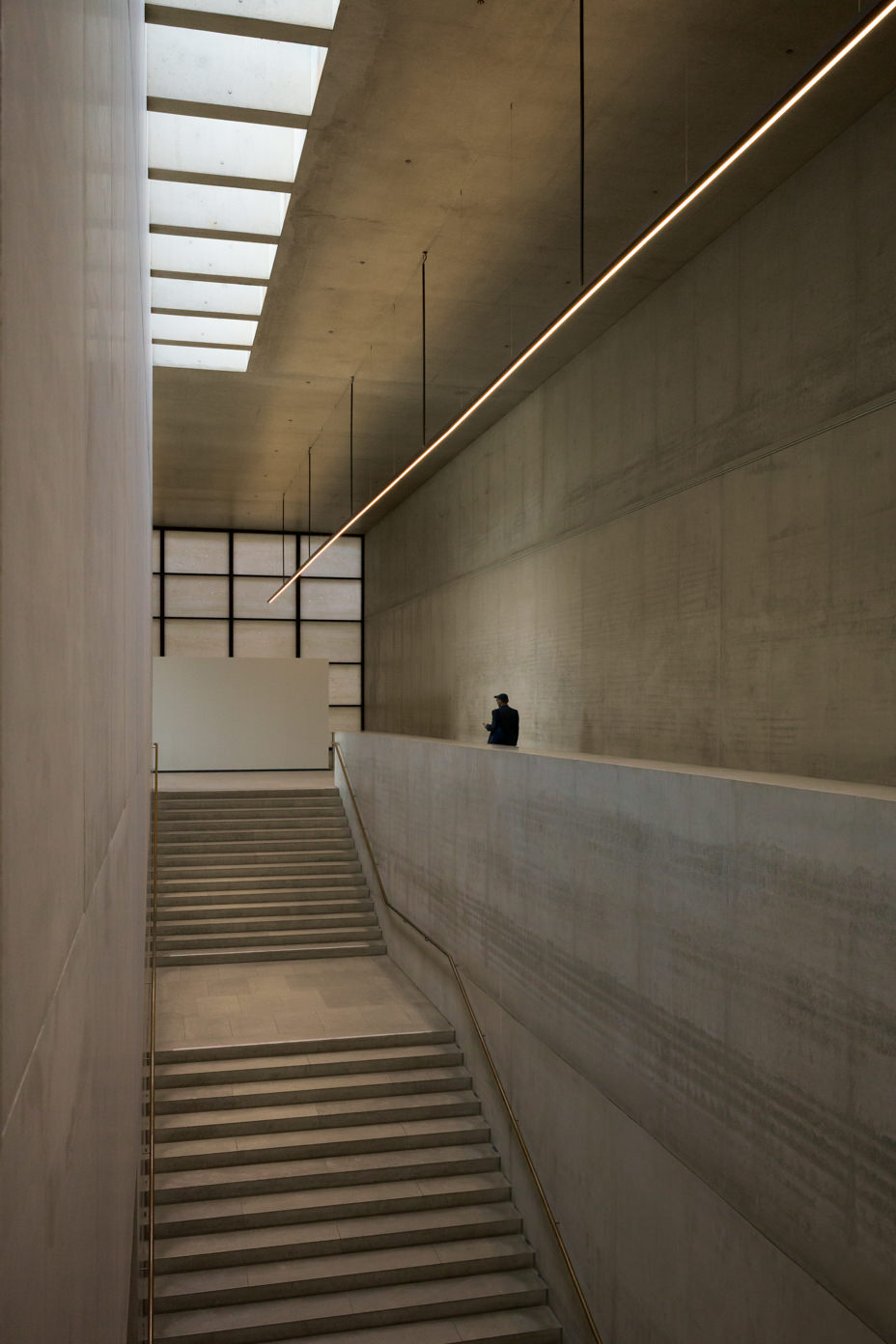 Tall concrete stairwell under a skylight, a thin line of light running diagonally; a lone man at the upper ledge, dwarfed by the space.