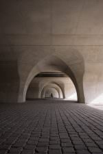 Row of pointed concrete arches beneath a bridge, cobblestones leading the eye to a small cyclist framed in the farthest arch.