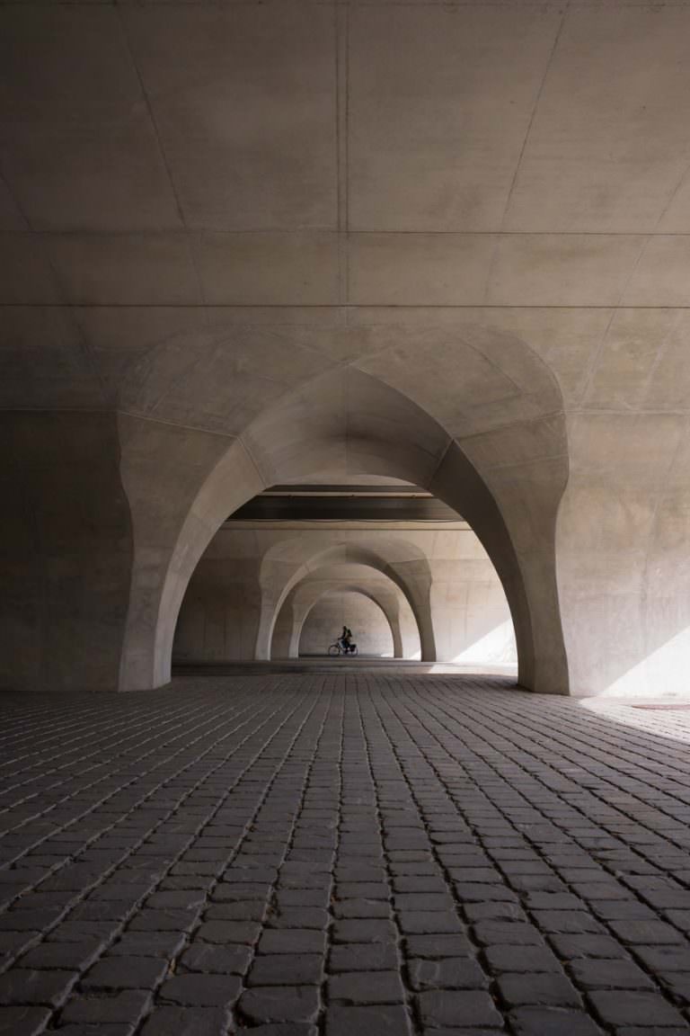 Row of pointed concrete arches beneath a bridge, cobblestones leading the eye to a small cyclist framed in the farthest arch.