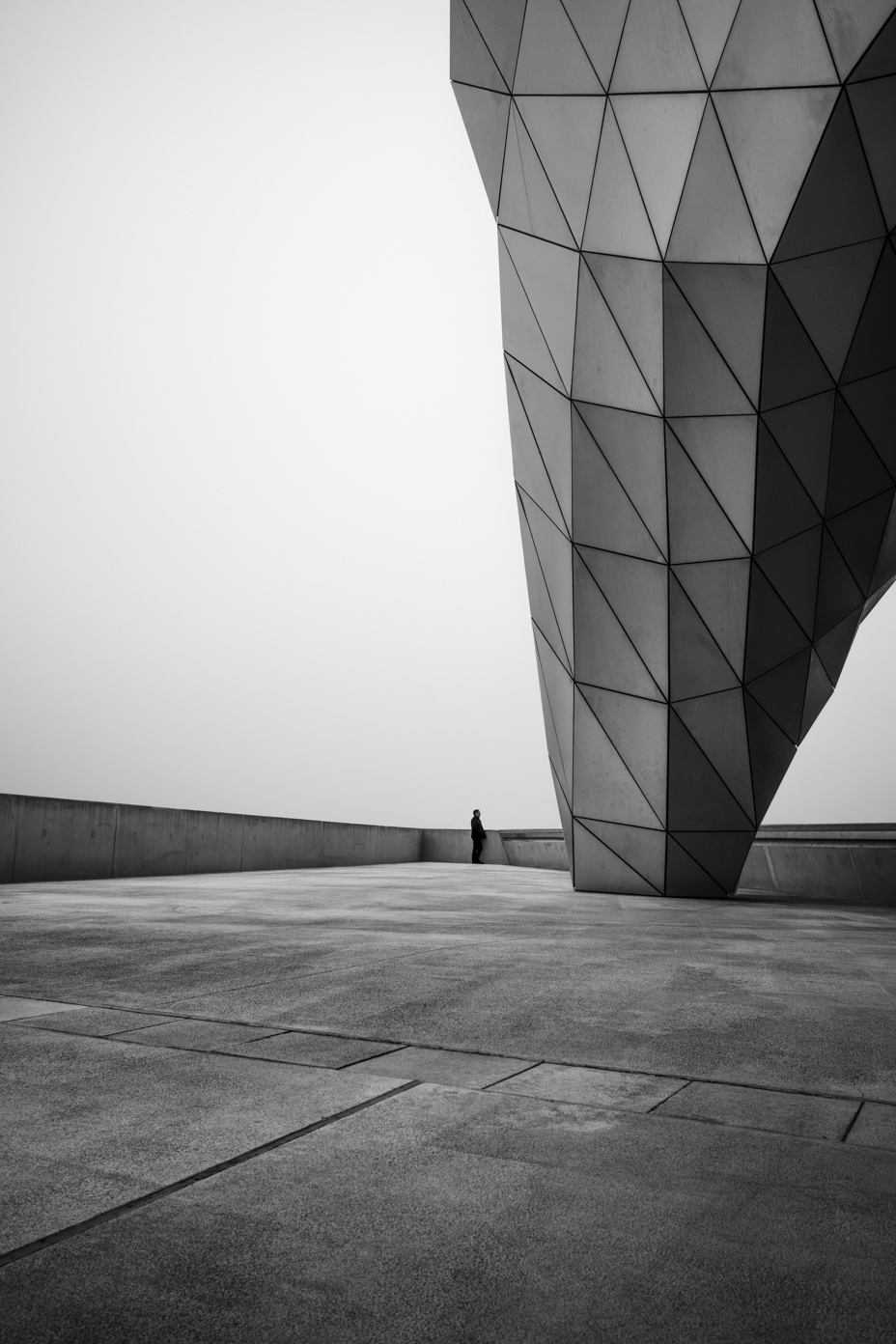 Black-and-white photo: A single tiny person stands at a concrete wall on an empty terrace in the fog. On the right, a huge building with a triangular facade towers into the frame.